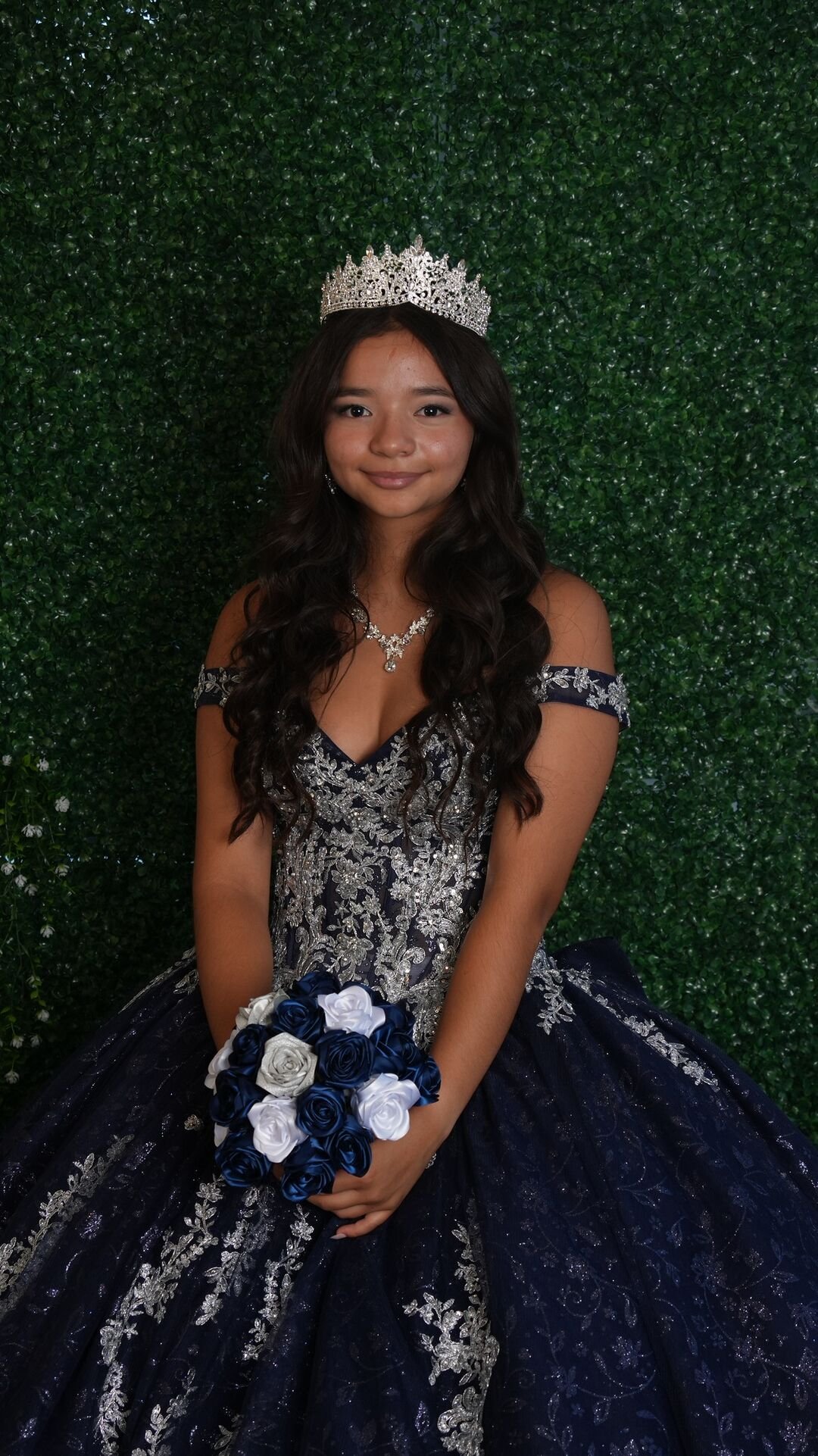 Full-length portrait of the quinceañera holding her bouquet in front of a greenery wall